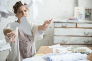 An annoyed woman is sitting at a desk and throwing her paperwork into the air in frustration.