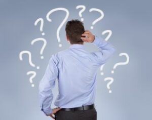 A person in business clothes in scratching their head while looking at a wall of question marks.