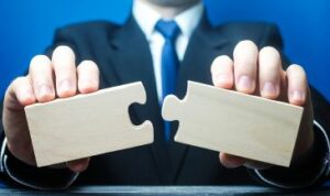A person in a dark business suit it holding out two large, wooden puzzle pieces and appears to be ready to connect them together.