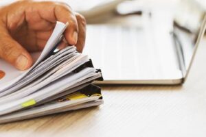 A person's hand is shown thumbing through a stack of documents on a desk.