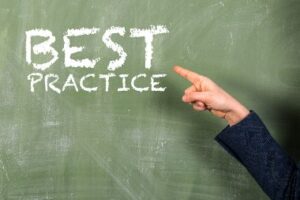 A person's hand is pointing to the words Best Practice that are written in chalk on a green chalkboard.