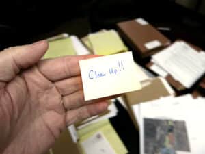 Hand holding a sticky note that reads "Clean Up" in front of a cluttered desk covered with stacks of papers and folders.